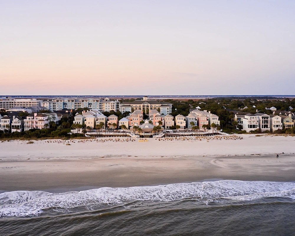 A row of beach houses along a sandy shore with gentle waves, calm sea, and a pastel sky at sunset, peaceful coastal scene.