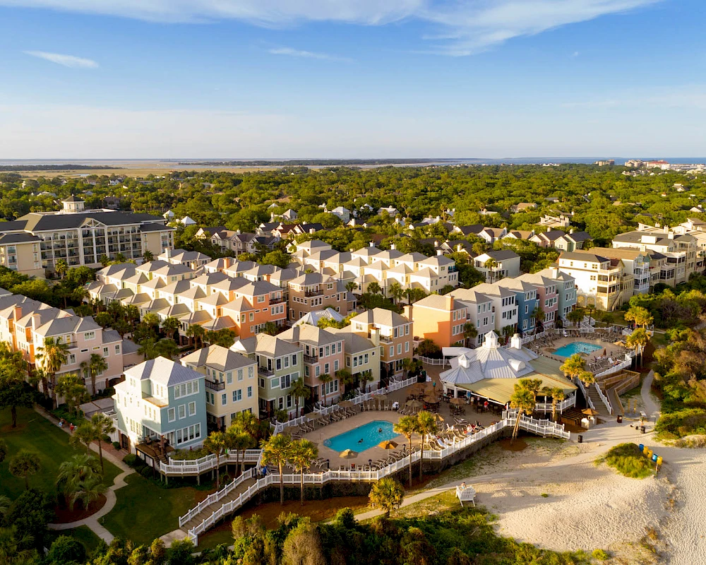 Aerial view of a beachfront residential community with pastel townhouses, a central courtyard, a pool, and sandy beaches meeting the sea.