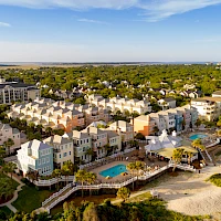 Aerial view of a beachfront residential community with pastel townhouses, a central courtyard, a pool, and sandy beaches meeting the sea.