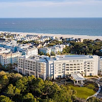 An aerial view shows a coastal resort with white apartment buildings, a sandy beach, and lush green trees in the foreground, ocean beyond.