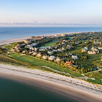 A coastal resort town with sandy beaches, grassy dunes, and clusters of houses along a curved shoreline by the calm sea.