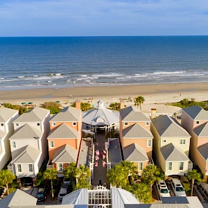 Colorful beachside cottages line up near the shore, with palm trees, a sandy beach, and calm waves beyond under a blue sky.
