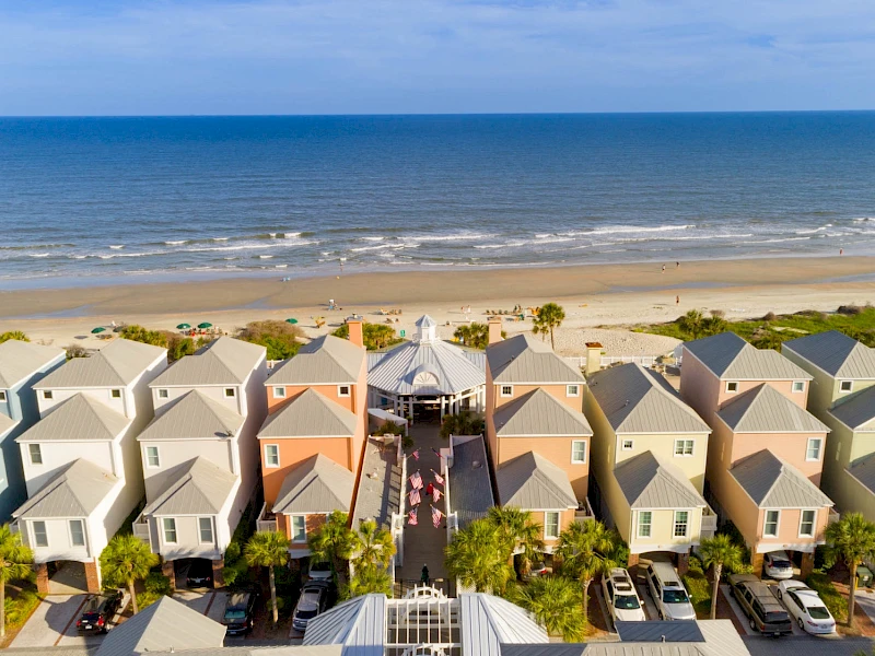 Colorful beachside cottages line up near the shore, with palm trees, a sandy beach, and calm waves beyond under a blue sky.