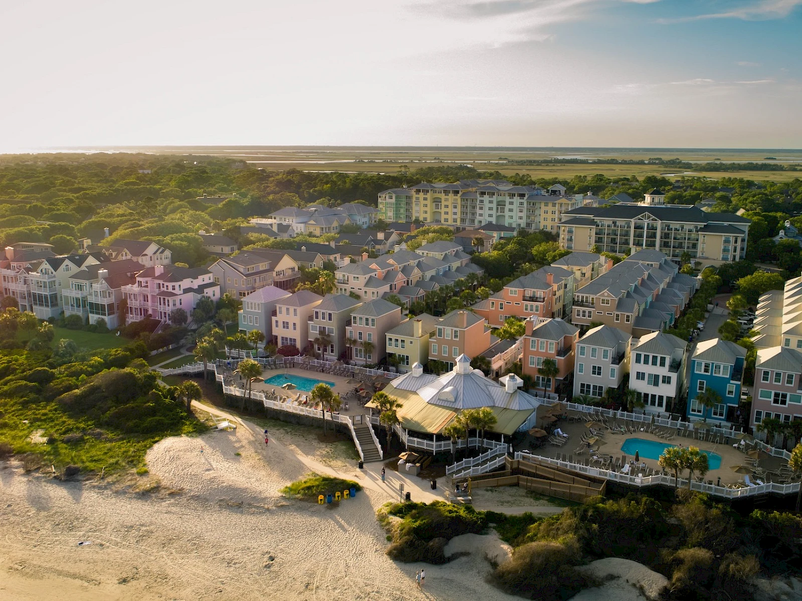 Coastal cliffside town with colorful multi-story beach houses, a row of pools and decks overlooking a sandy shoreline at sunset.