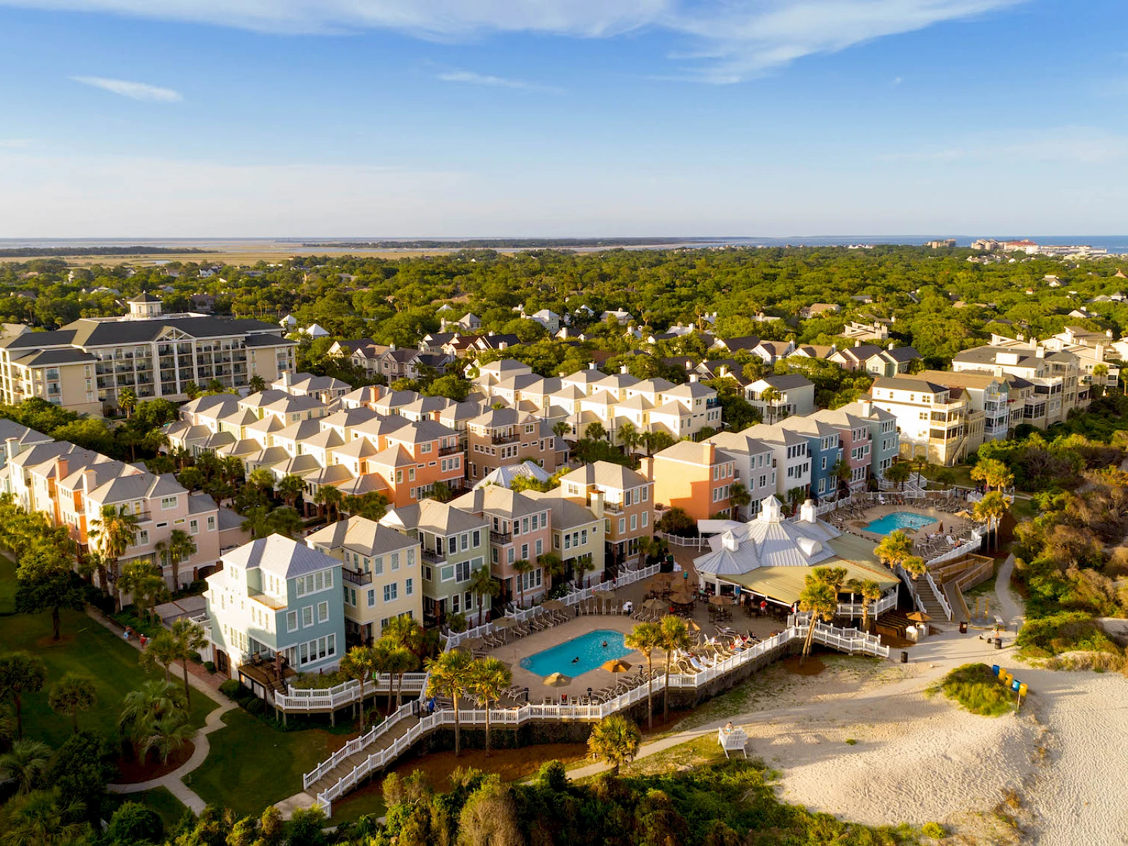 Aerial view of a coastal resort: pastel townhouses with a central pool, boardwalk, and sandy beach along the shoreline under a blue sky.