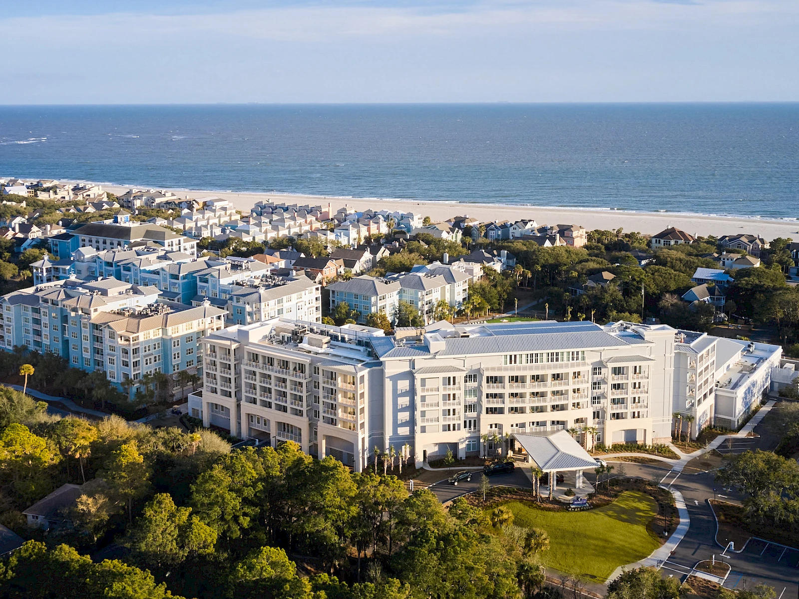 Aerial view of a coastal resort town with white apartment buildings, sandy beach, and lush green trees along the shore, by the ocean.