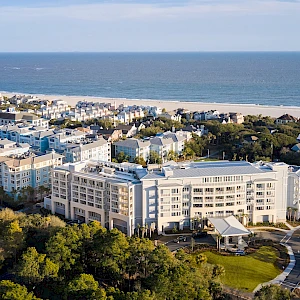 Aerial view of a coastal resort town with white apartment buildings, sandy beach, and lush green trees along the shore, by the ocean.