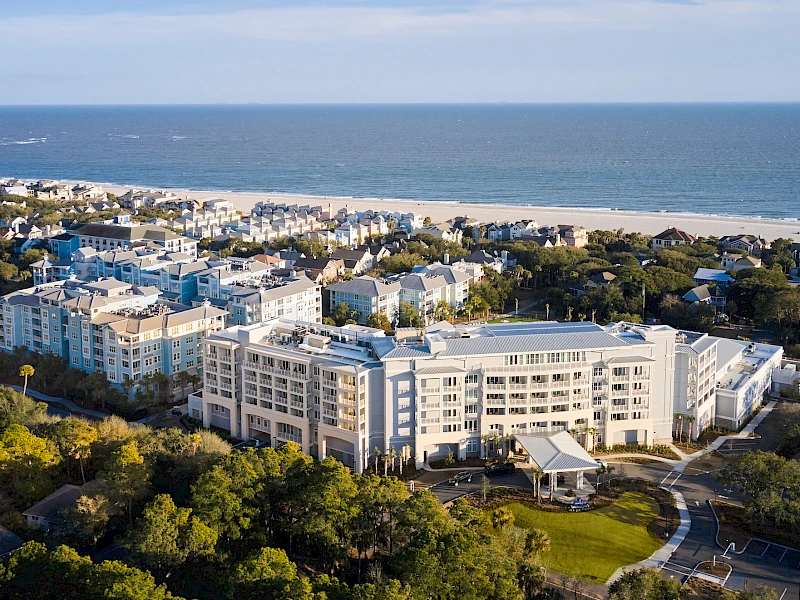 An aerial view of a coastal resort town with white multi-story buildings, a sandy beach, and lush green trees along the shoreline, near the sea.