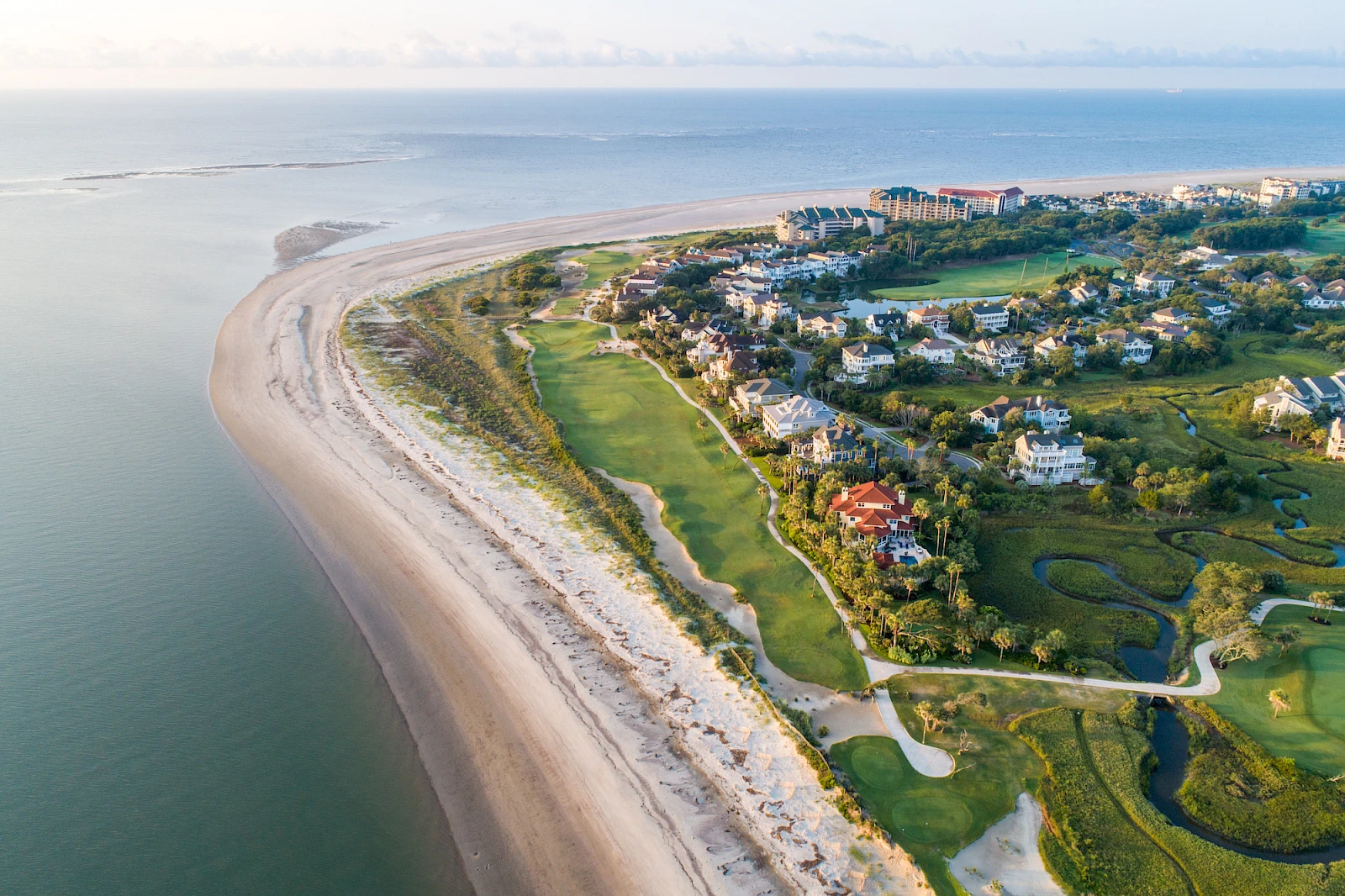 Aerial view of a coastal golf course along a sandy shoreline, with greens and fairways, water on one side, and residential buildings inland.