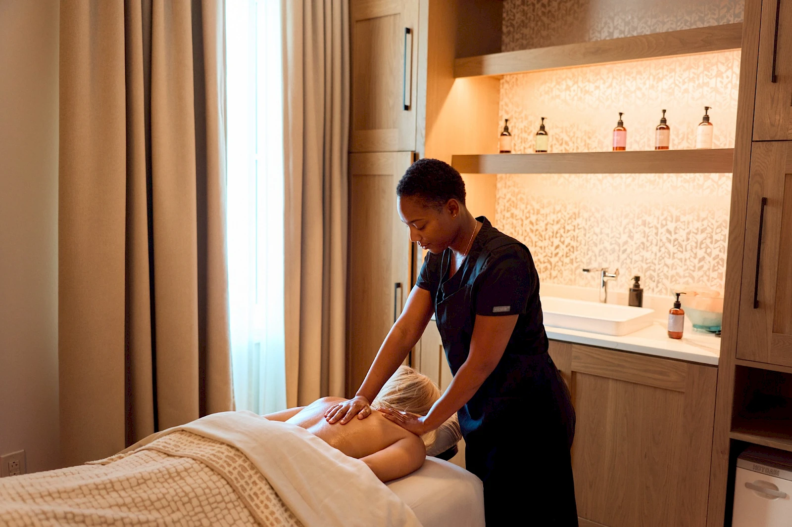 A masseuse gives a back massage to a person lying face down on a spa bed in a calm, well-lit treatment room.