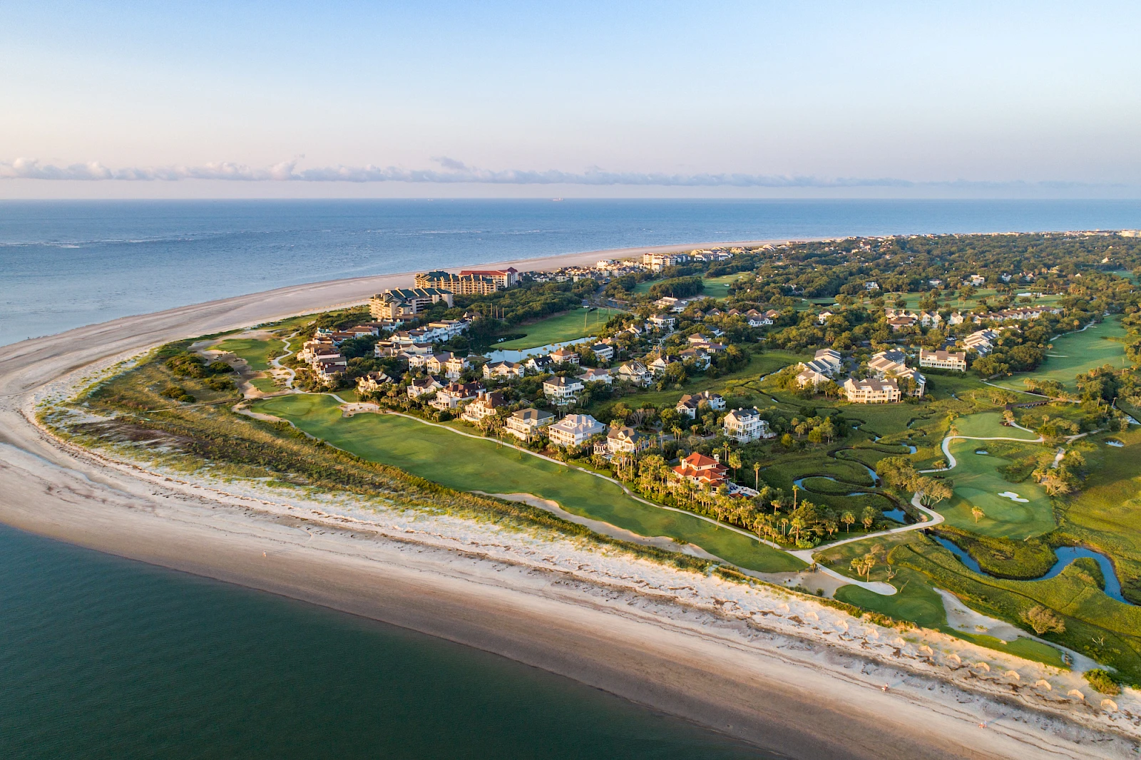Aerial view of a coastal resort town with sandy beach, green lawns, houses, and a winding waterway along the shoreline.