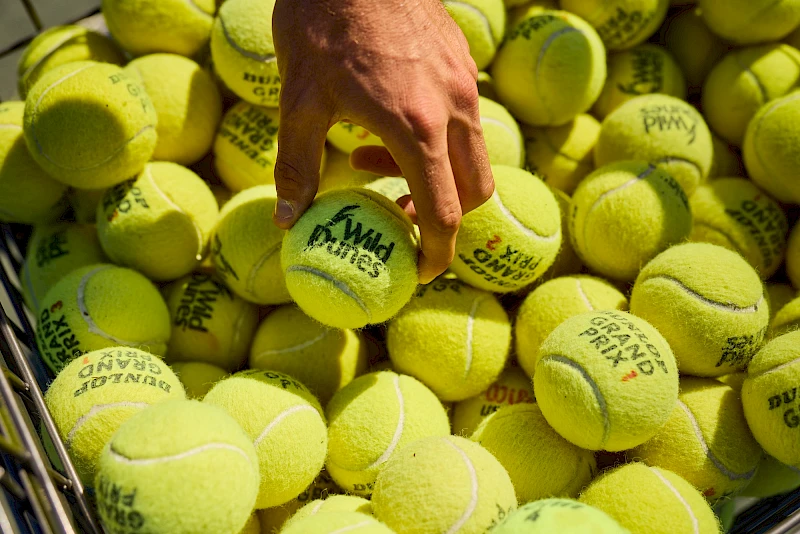A hand picks a bright yellow tennis ball from a pile of many similar balls.