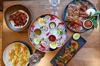 A spread of seafood and tapas: oysters on ice with lemon wedges, charred seafood bites, crispy toasts, smoked salmon platter, and a dessert bite platter, all on a wooden table.