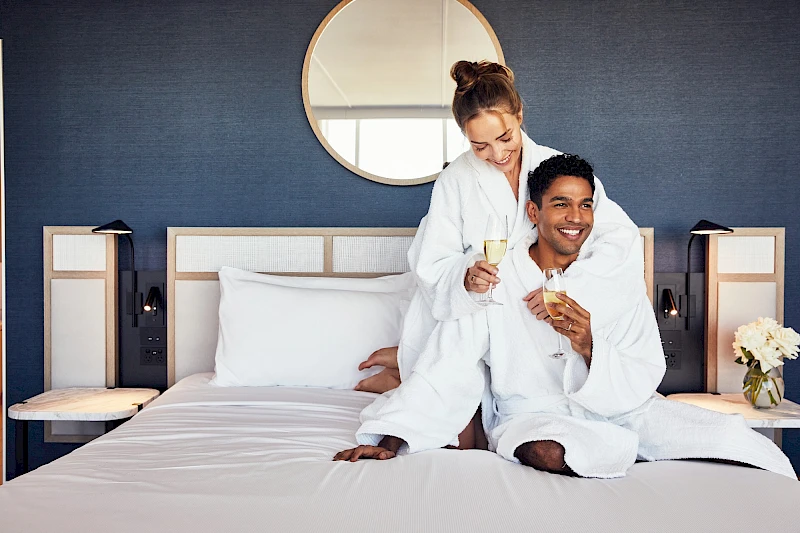 Two people in white robes toast with champagne on a bed, smiling in a stylish hotel room.