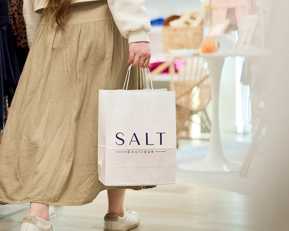 A woman walks in a boutique carrying a white bag that says “SALT,” wearing a beige skirt, white top, and sneakers, storefronts and shoppers in the background.