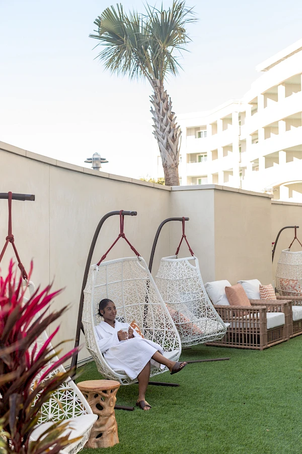 A person relaxing in a hanging chair by a poolside garden with palm trees and modern white buildings in the background.
