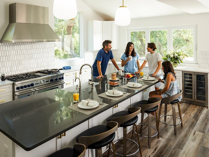 A modern, bright kitchen with a long island counter, bar stools, stainless steel appliances, and a family chatting while prepping food.