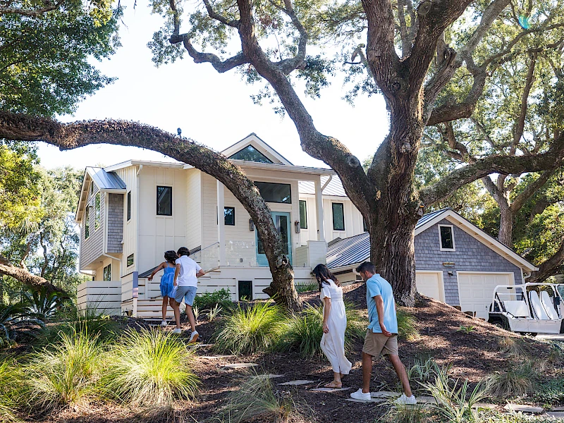 A family visits a white, two-story house with a wraparound porch, walking along a tree-lined path on a sunny day.