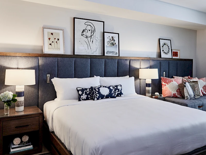 A neatly made king bed with white linens, dark upholstered headboard, two nightstands, lamps, and framed art above the bed in a stylish hotel-like bedroom.