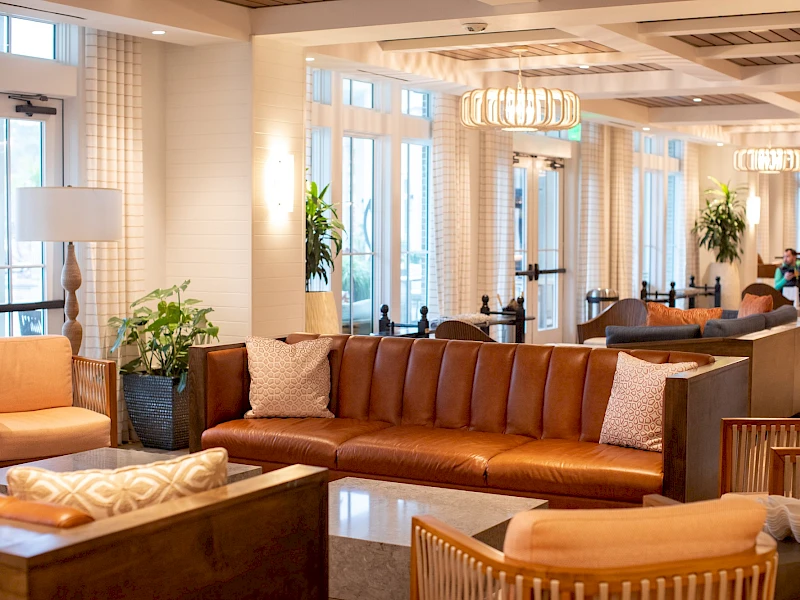 A bright hotel lobby with tan leather sofas, wooden coffee tables, plants, and large windows; people chatting in the background.