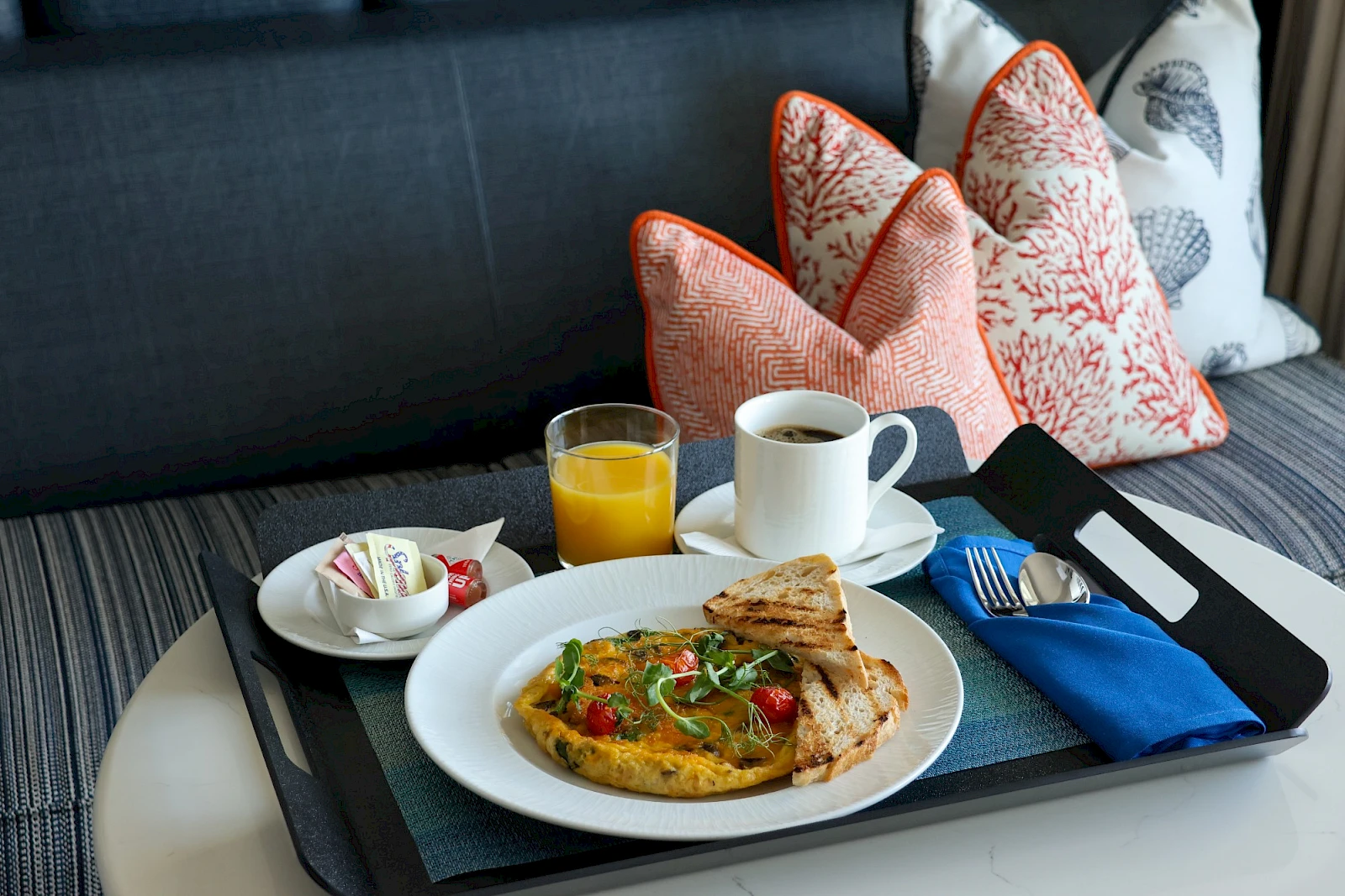 A breakfast tray with a plate of food, a glass of orange juice, coffee, toast, and a small side plate on a cushioned seat with patterned pillows.