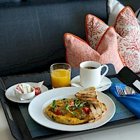 A breakfast tray with a plate of food, a glass of orange juice, coffee, toast, and a small side plate on a cushioned seat with patterned pillows.
