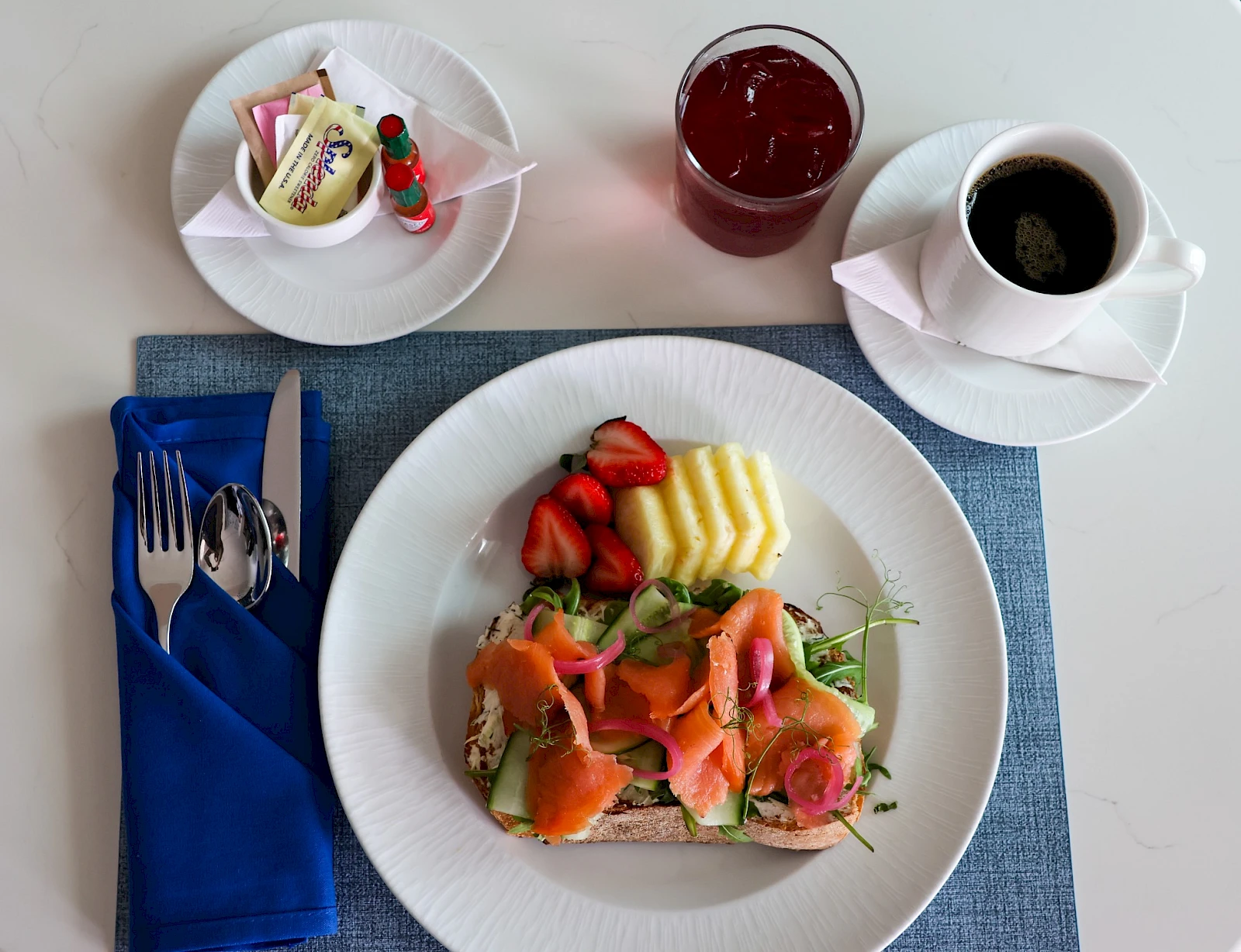 A breakfast/meal setting with a plate of smoked salmon and toppings, pineapple and strawberries, a blue napkin, a cup of coffee, and a glass of juice on a light table.