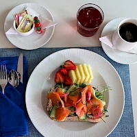 A breakfast/meal setting with a plate of smoked salmon and toppings, pineapple and strawberries, a blue napkin, a cup of coffee, and a glass of juice on a light table.
