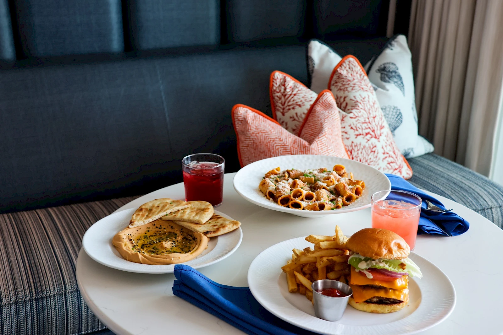 A restaurant table with pancakes, pasta, a burger with fries, and drinks on a cozy couch setup, ready for a hearty meal tonight.