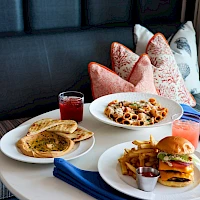 A restaurant table with pancakes, pasta, a burger with fries, and drinks on a cozy couch setup, ready for a hearty meal tonight.