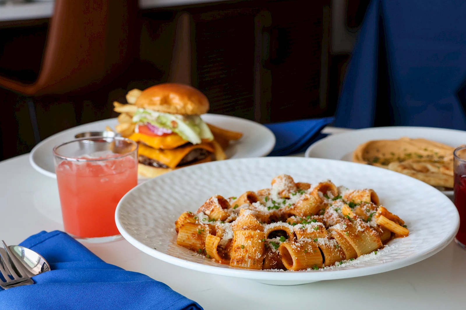 A plated pasta dish with croutons, cheese, and greens sits front and center, a burger and pink drink visible in the background.