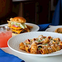 A plated pasta dish with croutons, cheese, and greens sits front and center, a burger and pink drink visible in the background.
