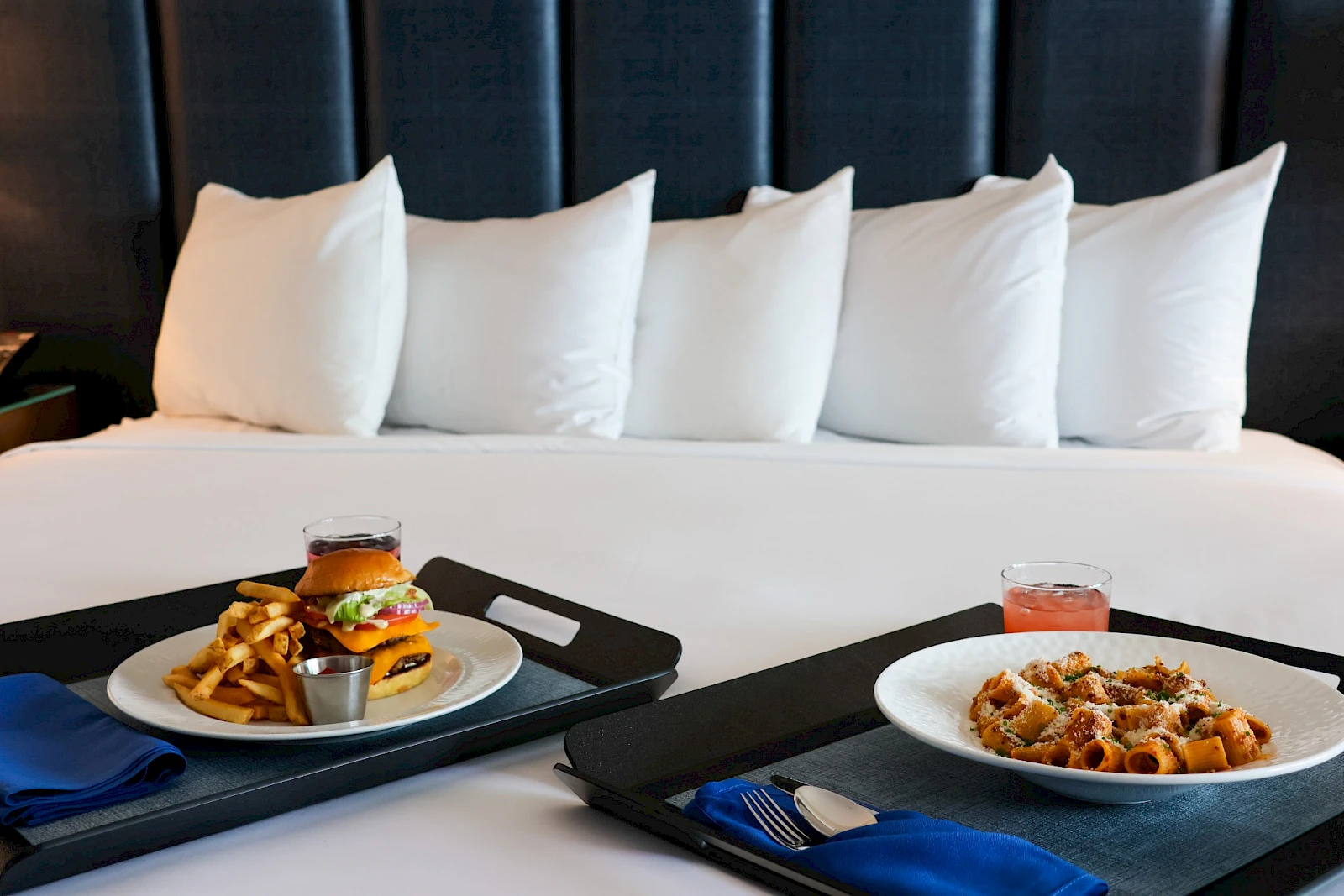 Two plated meals on a hotel bed with pillows; burger and fries on one tray, pasta on the other, plus drinks and utensils.