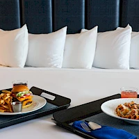 Two plated meals on a hotel bed with pillows; burger and fries on one tray, pasta on the other, plus drinks and utensils.