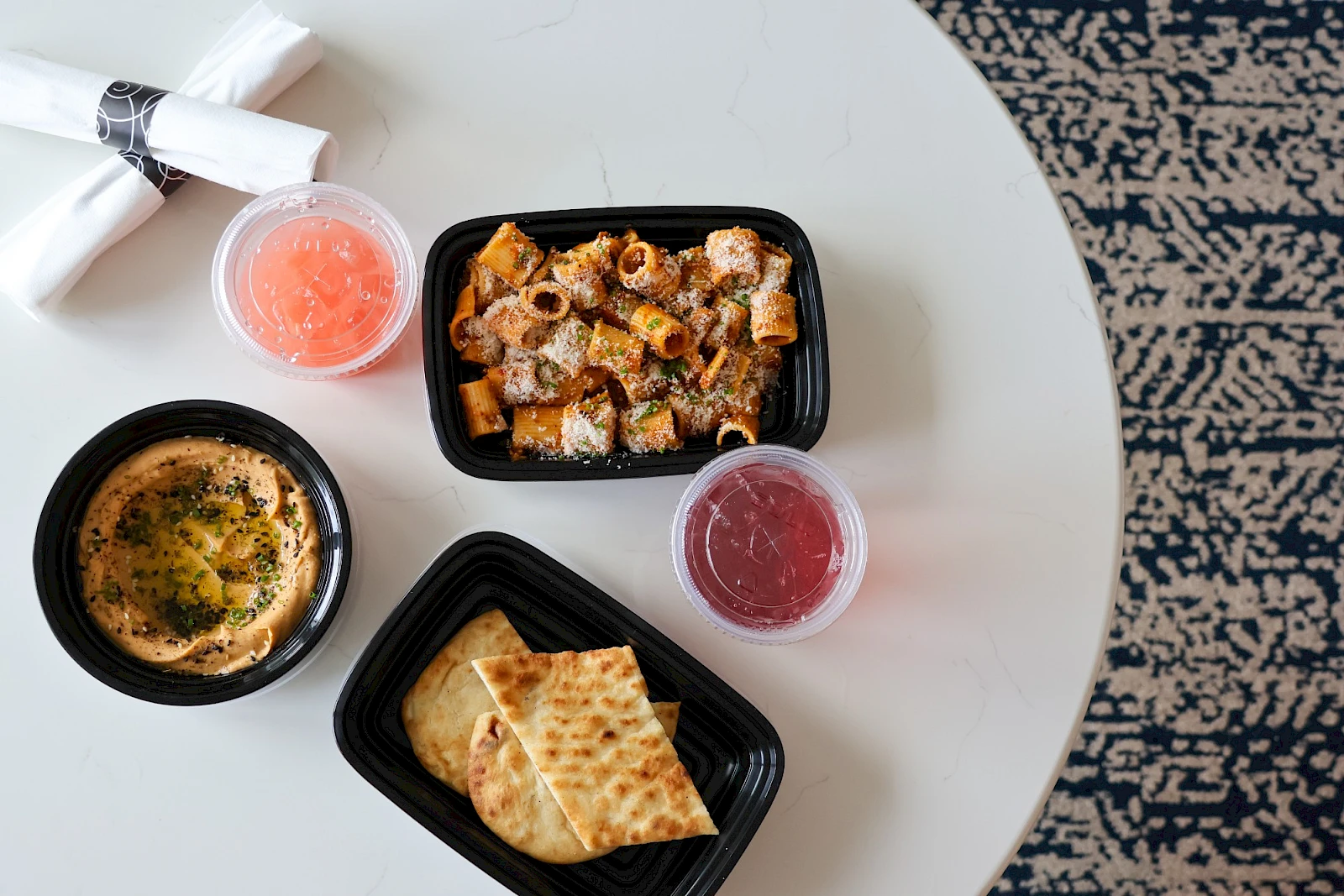 A round table with takeout containers: creamy dip, spiced meat mix, toasted pita, and two fruit/tea cups, on a patterned rug.