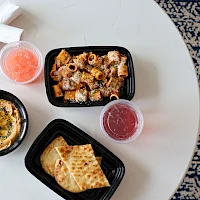A round table with takeout containers: creamy dip, spiced meat mix, toasted pita, and two fruit/tea cups, on a patterned rug.