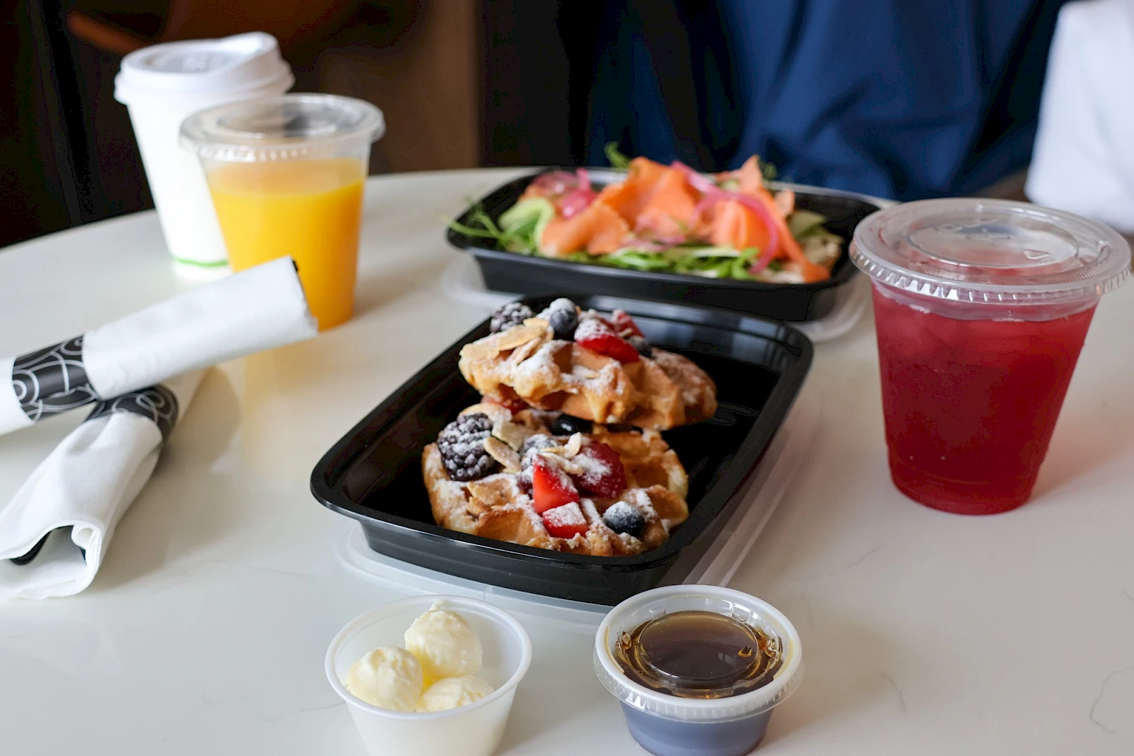 A breakfast spread with waffles topped with berries, a side salad, fruit juice and a red drink, a small cup with butter and syrup, plus napkins on a white table.