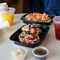 A breakfast spread with waffles topped with berries, a side salad, fruit juice and a red drink, a small cup with butter and syrup, plus napkins on a white table.