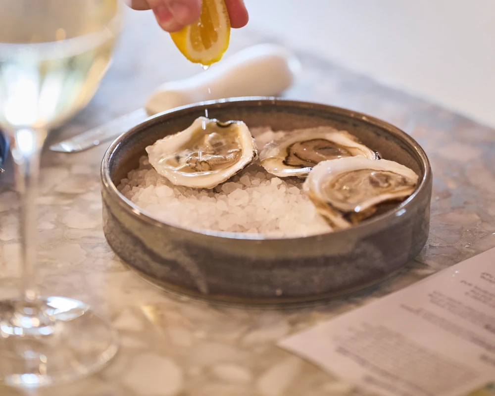 A hand sprinkles lemon over a plate of oysters on ice, with a wine glass nearby, on a shiny table.
