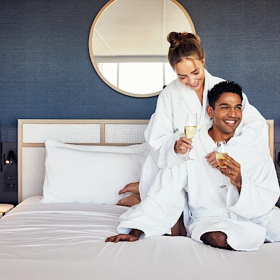 Two smiling people in bathrobes celebrate with champagne on a hotel bed, enjoying a cozy, celebratory moment together.