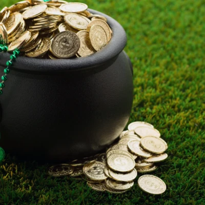 A black pot overflowing with coins sits on green grass, with a green shamrock and bead necklace draped over the side, on St. Patrick’s Day vibes.