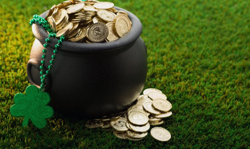 A black pot overflowing with coins sits on green grass, with a green shamrock and bead necklace draped over the side, on St. Patrick’s Day vibes.