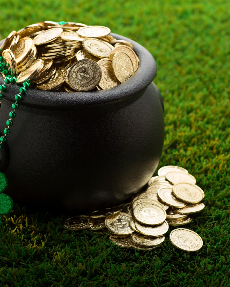 A pot of coins on green grass with a green shamrock bead bracelet, overflowing with shiny coins and a few coins spilled nearby.