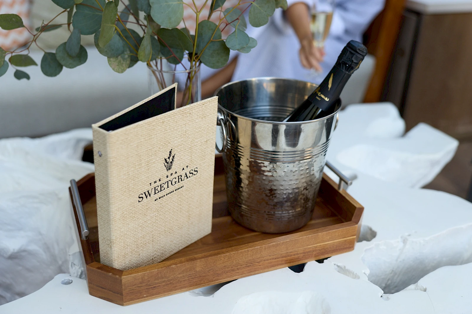A wooden tray with a metal ice bucket holding a bottle, a small menu/book, and eucalyptus leaves on a cozy bed setup.
