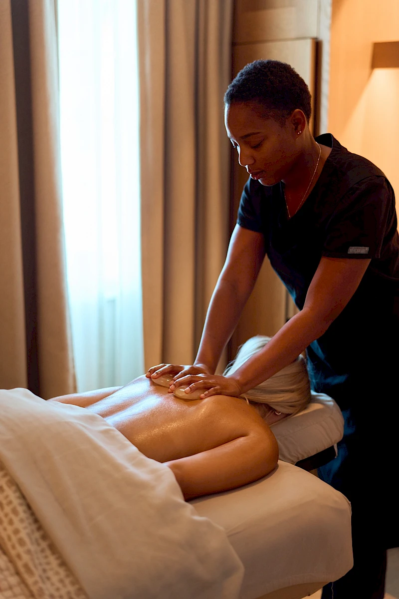 A massage therapist applies pressure to a client's lower back while the client lies face down on a bed, towel draped, in a calm, softly lit room.