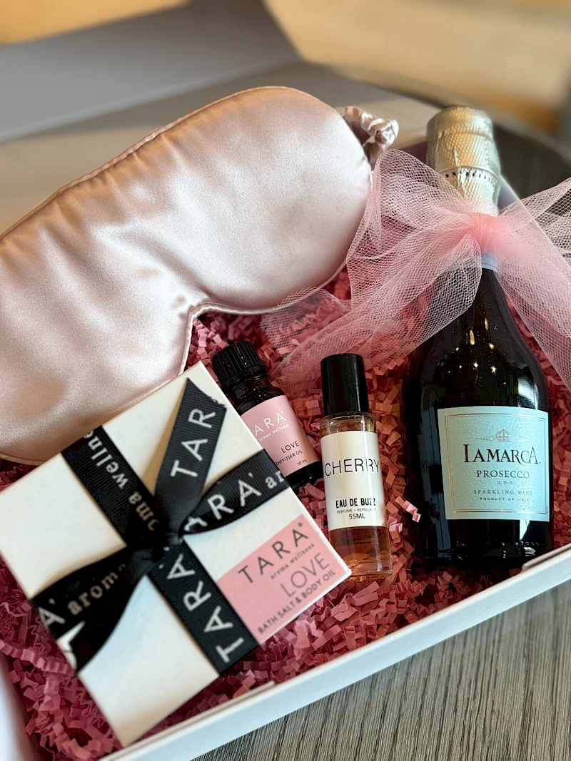 A beauty gift box with small perfume bottles, a pink-tinted bottle, a champagne-looking bottle, and black-and-white labeled items, all on pink filler.