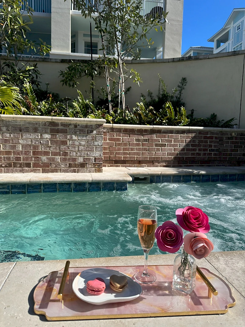 A chic poolside setup: a tray with a plate of dessert, a glass of champagne, and a small vase of pink roses overlooking a sunny pool.
