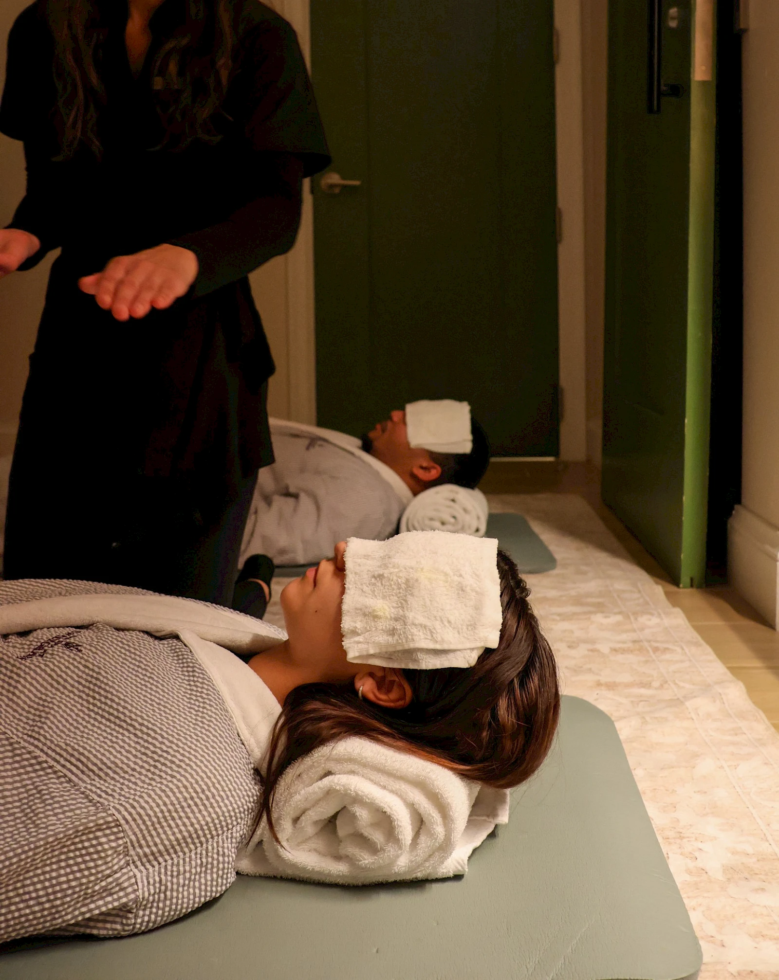 People receiving spa or massage therapy; two clients lie face down with towels on their heads, a practitioner stands nearby.