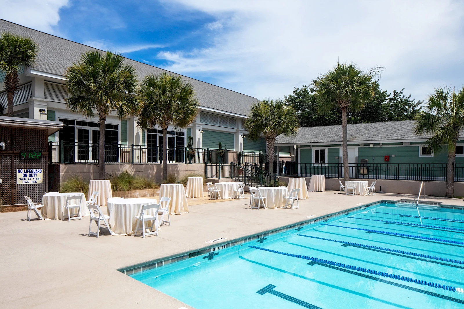 Outdoor hotel pool area with clear blue water, sun loungers and small round tables, palm trees, and a light building in the background.