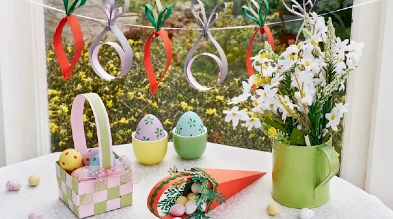 A festive Easter setup: pastel eggs, a basket with small toys, decorative rings hanging above, a bouquet in a green jug, and spring colors. ending with a period.